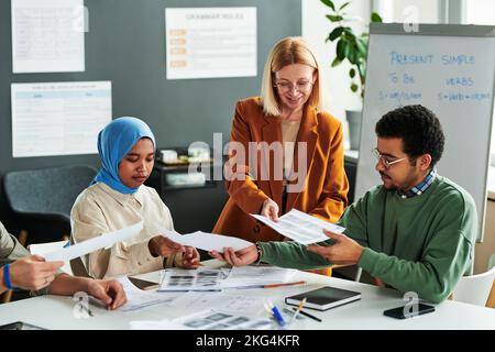 Eine Gruppe interkultureller Schüler, die Grammatiktests machen, während der Lehrer ihnen während des Unterrichts der englischen Sprache Papiere übergibt Stockfoto