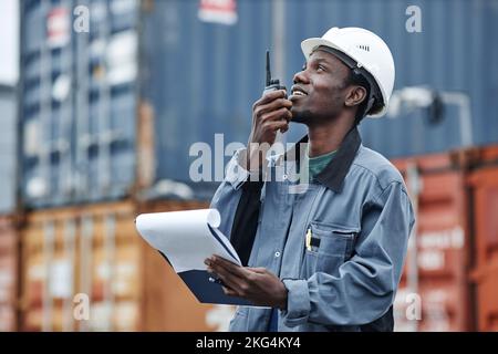 Seitenansicht Porträt eines jungen schwarzen Mannes mit Walkie-Talkie und Blick nach oben, während er an den Schiffsanlegestellen mit Containern arbeitete Stockfoto