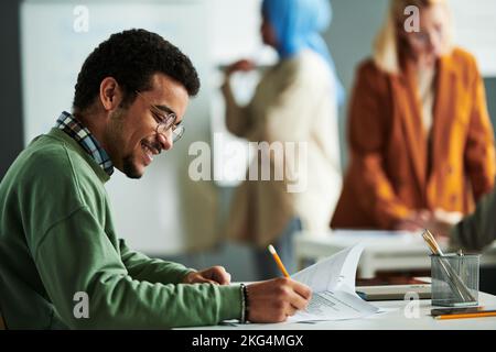 Seitenansicht eines glücklichen Studenten aus dem Nahen Osten, der im Klassenzimmer am Schreibtisch einen Grammatiktest durchführt und dabei vor die richtigen Antworten tickt Stockfoto
