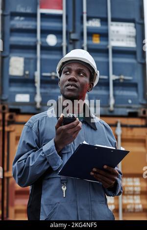 Vertikale Taille oben Porträt eines schwarzen männlichen Arbeiters trägt Hardhut in Schiffsanlegestellen gegen Container Stockfoto