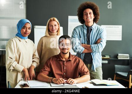 Eine Gruppe multikultureller Schüler und reifer blonder Lehrer in lässiger Kleidung, die vor der Kamera mit Whiteboard hinter dem Schreibtisch stehen Stockfoto
