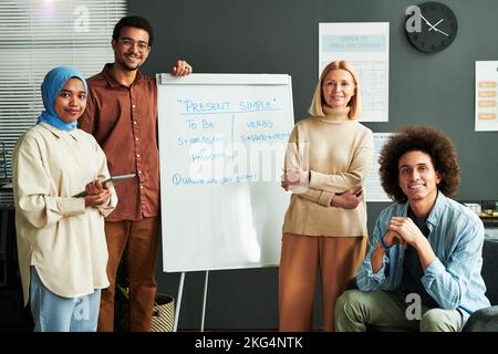 Mehrere glückliche junge interkulturelle Studenten und reife blonde Lehrer rund Whiteboard mit Erklärung der Present Simple Tense Stockfoto