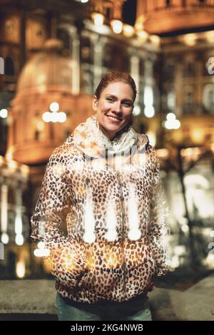 Glückliche junge Rothaarige Stadtfrau im Leopardenmuster Mantel im Winter toothy Lächeln draußen in der Nacht Stockfoto
