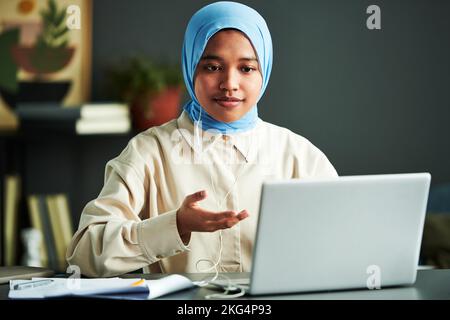 Junge selbstbewusste muslimische Frau in Hijab, die am Webinar teilnimmt, während sie zu Hause vor dem Laptop am Schreibtisch sitzt und etwas erklärt Stockfoto