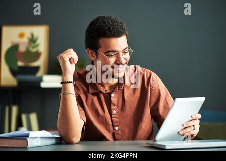 Junger lächelnder männlicher Student des Online-Studiengangs beim Betrachten des Tablet-Bildschirms beim Sitzen am Schreibtisch und beim Betrachten der Videostunde Stockfoto