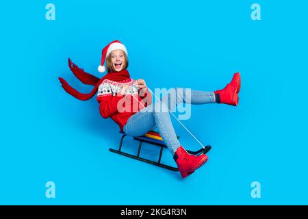 Foto in voller Länge von einem flippigen, beeindruckenden Schulmädchen in roter Weihnachtsdekoration Pullover Schlittenfahrten mit isoliertem blauem Hintergrund Stockfoto