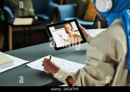 Konzentrieren Sie sich auf den Tablet-Bildschirm, wobei der blonde Lehrer selbstbewusst auf das Whiteboard zeigt, während er dem Online-Publikum die englische Grammatik erklärt Stockfoto