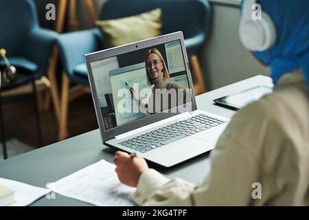 Selbstbewusster blonder Lehrer auf dem Laptop-Bildschirm, der auf das Whiteboard zeigt, während er Online-Schülern die neue englische Grammatik erklärt Stockfoto