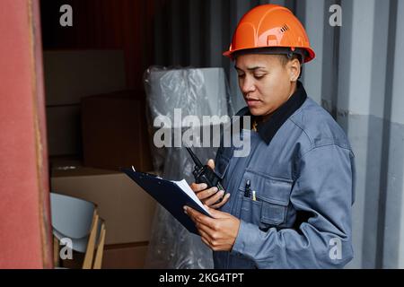 Seitenansicht Porträt einer multiethnischen Arbeitnehmerin, die einen Hardhut trägt, während sie die Container am Schiffsanleger überprüft und die Zwischenablage hält Stockfoto