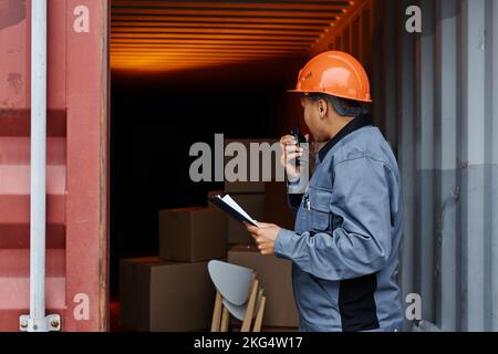 Seitenansicht Porträt einer Arbeiterin, die einen Hardhut trägt und mit dem Radio spricht, während sie die Container am Schiffsanleger überprüft und Platz kopiert Stockfoto