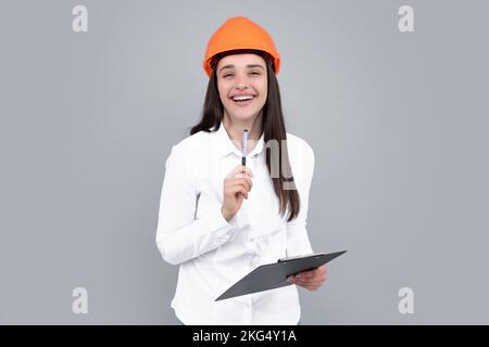 Lächelnde Frau im Schutzhelm mit Klemmbrett isoliert auf grauem Hintergrund. Junge Frau, Bauleiterin. Architektin, weibliche Mitarbeiterin Stockfoto