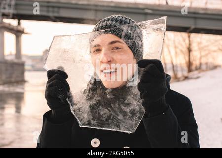 Teenager-Junge hält ein Stück Eis vor seinem Gesicht und schaut durch Eis, während er bei kaltem, frostigem Wetter draußen steht Stockfoto