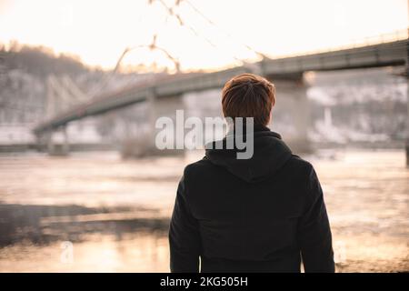 Rückblick auf einen rücksichtsvollen jungen Mann, der im Winter im Freien auf den Fluss blickt Stockfoto