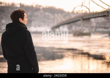 Rückblick auf einen nachdenklichen jungen Mann, der im Winter am Fluss steht Stockfoto