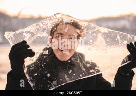 Ein fröhlicher Teenager, der bei kaltem, frostigem Wetter im Winter durch ein Stück Eis schaut und es dabei im Freien hält Stockfoto