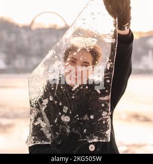 Glücklicher Teenager, der bei kaltem, frostigem Wetter im Winter durch ein Stück Eis schaute und es im Freien hielt Stockfoto