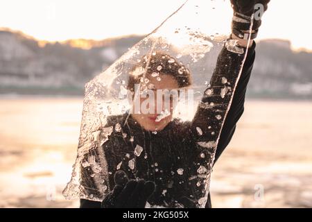 Teenager-Junge, der bei kaltem, frostigem Wetter im Winter durch ein Stück Eis schaut und es im Freien hält Stockfoto