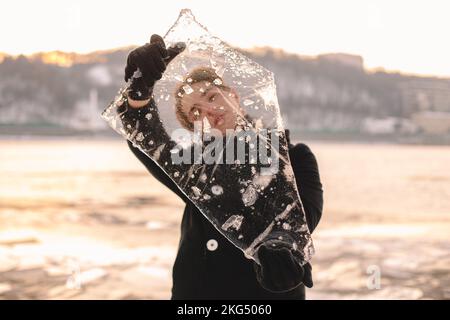 Teenager-Junge, der bei kaltem, frostigem Wetter im Winter durch ein Stück Eis schaut und es im Freien hält Stockfoto