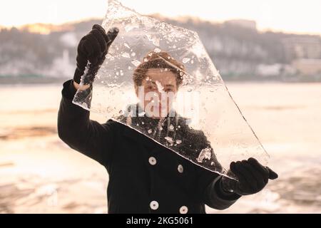 Teenager-Junge hält ein Stück Eis vor seinem Gesicht und schaut durch Eis, während er bei kaltem, frostigem Wetter draußen steht Stockfoto