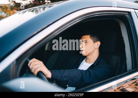 Ein junger Mann am Steuer eines Autos wird kabellose Kopfhörer benutzen Stockfoto