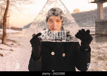 Teenager-Junge hält ein Stück Eis vor seinem Gesicht und schaut durch Eis, während er bei kaltem, frostigem Wetter draußen steht Stockfoto