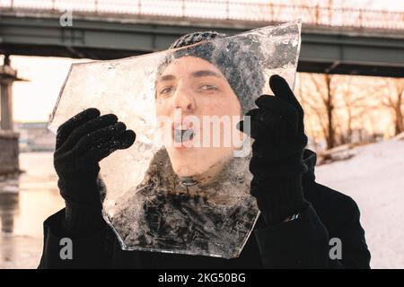 Teenager-Junge, der bei kaltem, frostigem Wetter im Winter durch ein Stück Eis schaut und es im Freien hält Stockfoto