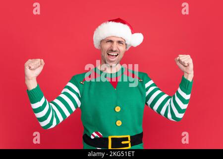 Spüren Sie den Erfolg. Fröhlicher schreiender Mann in Elfenkostüm. der weihnachtsmann mit dem weihnachtsmann-Hut Stockfoto