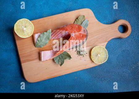 Ein Stück Fisch liegt auf einem Brett auf blauem Hintergrund. In der Nähe liegt eine halbe Zitrone und ein Lorbeerblatt. Stockfoto