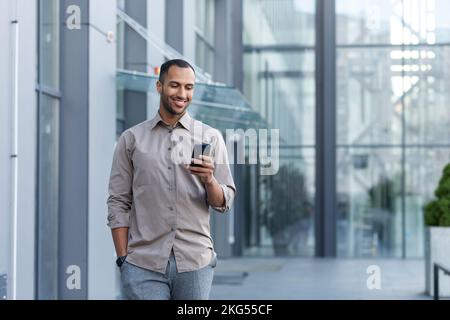 Ein junger, gutaussehender Hispanic, Afroamerikaner läuft die Straße runter in der Nähe eines Bürozentrums. Er hält ein Telefon in der linken Hand, seine rechte Hand in der Tasche. Benutzt, liest die Nachrichten, lächelt. Stockfoto