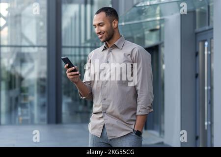 Ein junger, gutaussehender, lateinamerikanischer Mann steht auf der Straße in der Nähe eines Bürozentrums. Er hält das Telefon in der Hand, benutzt es, wählt, lächelt. Stockfoto