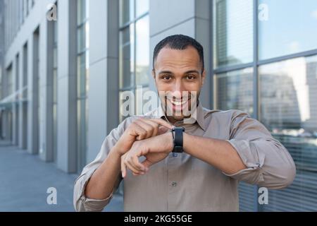 Junger, gutaussehender, afroamerikanischer Geschäftsmann, der auf Smartwatch spricht. Es steht auf der Straße in der Nähe des Bürozentrums. Er sieht in die Kamera und lächelt. Stockfoto