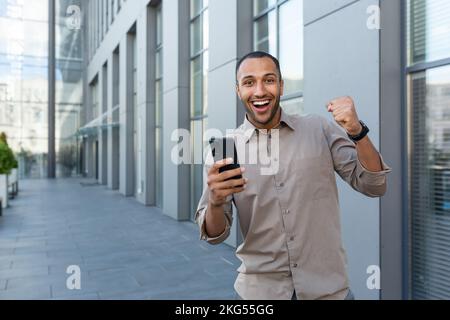 Junger gutaussehender Hispanic, Afroamerikaner, der ein Handy in der Hand hält, Online-Spiele spielt, ja mit der Hand zeigt. Er schaut in die Kamera, freut sich über den Sieg, ich werde gewinnen. Stockfoto