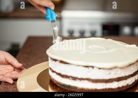 Eine Frau, die in der Küche die Kanten eines Schokoladenkuchens mit Schlagsahne und einem Teigschaber glättet Stockfoto