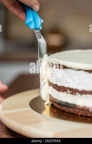 Eine vertikale Aufnahme einer Frau, die in der Küche die Kanten eines Schokoladenkuchens mit Schlagsahne und einem Spachtel glätten soll Stockfoto