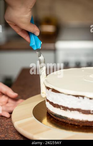 Eine vertikale Aufnahme einer Frau, die in der Küche die Kanten eines Schokoladenkuchens mit Schlagsahne und einem Spachtel glätten soll Stockfoto