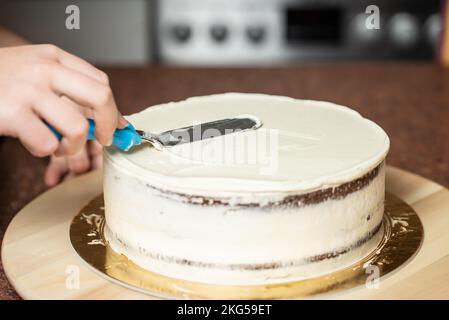 Eine Frau, die in der Küche die Kanten eines Schokoladenkuchens mit Schlagsahne und einem Teigschaber glättet Stockfoto
