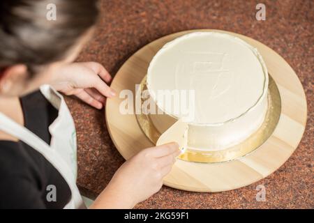 Eine Frau, die in der Küche die Kanten eines mit Schlagsahne überzogenen Schokoladenkuchens mit einem glatteren Werkzeug glätten sollte Stockfoto