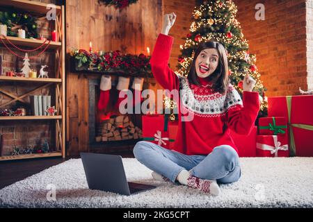 Großes Foto von einem entzückten, überfreulichen Mädchen, das auf dem Teppich sitzt. Verwenden Sie ein Netbook, um die Fäuste zu heben, Glück Girlande Lichter drinnen Stockfoto