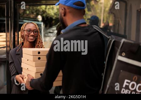 Lieferung von Geschäftsessen, fröhlicher Kunde mit Pizzakartons, lächelnde Frau, die Bestellung vom Kurier in der Nähe des Bürogebäudes erhält. afroamerikanischer Angestellter, der Essenspakete stapelt Stockfoto