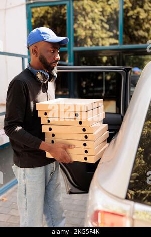 Lieferer, der Pizza mit dem Auto ins Büro brachte, Seitenansicht, afroamerikanischer Mann, der Kartonstapel hat. Mitarbeiter des Lunch-Service, der Fastfood liefert, steht in der Nähe des Firmengebäudes im Freien Stockfoto