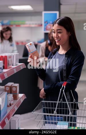 Lächelnder asiatischer Apothekenkunde Warenkorb mit Blick auf Regale mit Gesundheitsprodukten Ergänzungen kaufen. Kunde wählt Vitamine für das Immunsystem im pharmazeutischen Shop Stockfoto