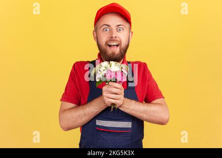 Porträt eines verblüfften, verblüfften Auslieferers mit Blumenstrauß, der mit überraschtem Gesichtsausdruck auf die Kamera schaut, blaue Uniform und rote Mütze trägt. Innenstudio-Aufnahme isoliert auf gelbem Hintergrund. Stockfoto