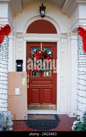 Hübsche Wohnveranda, dekoriert für Weihnachten mit Kranzleuchten und Schleifen und einem großen braunen Paket, das gegen Wand an Seite gewickelt geliefert wird Stockfoto