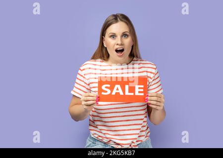 Porträt einer überraschten, überraschten Frau, die ein gestreiftes T-Shirt trägt, mit einer Verkaufskarte in den Händen steht und mit offenem Mund und großen Augen in die Kamera schaut. Studio-Aufnahme im Innenbereich isoliert auf lila Hintergrund Stockfoto