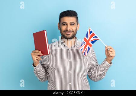 Porträt eines attraktiven jungen Erwachsenen mit Buch und britischer Flagge, Bildung im Ausland, Blick in die Kamera, mit gestreiftem Hemd. Innenstudio, isoliert auf blauem Hintergrund. Stockfoto