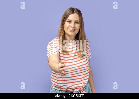 Darf ich mich vorstellen? Porträt einer freundlichen blonden Frau, die ein gestreiftes T-Shirt trägt, die Hand schüttelt und die Gäste mit einem zahnigen Lächeln begrüßt. Studio-Aufnahme im Innenbereich isoliert auf lila Hintergrund. Stockfoto