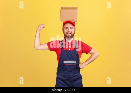 Portrait eines starken Kuriers in blauer Uniform, der mit einem Karton auf dem Kopf stand, in die Kamera schaute, den erhobenen Arm seiner Macht zeigte. Innenstudio-Aufnahme isoliert auf gelbem Hintergrund. Stockfoto