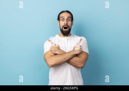 Porträt eines überraschten Mannes mit Bart und weißem T-Shirt, der mit gekreuzten Händen auf den Kopierbereich auf beiden Seiten zeigt. Innenstudio, isoliert auf blauem Hintergrund. Stockfoto