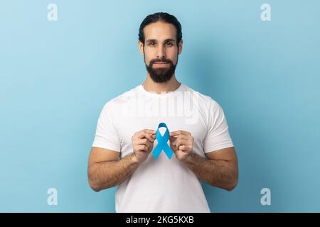 Portrait eines seriösen verantwortlichen Mannes mit Bart trägt weißes T-Shirt mit blauem Bewusstsein, Krankheitssymbol, schaut in die Kamera, Unterstützung. Innenstudio, isoliert auf blauem Hintergrund. Stockfoto