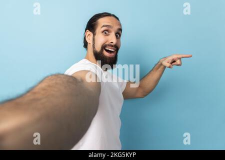 Porträt eines aufgeregten Mannes, der ein weißes T-Shirt trägt, mit positivem Ausdruck in die Kamera schaut, auf den Kopierbereich zeigt, auf die POV, auf die Perspektive des Fotos. Innenstudio, isoliert auf blauem Hintergrund. Stockfoto